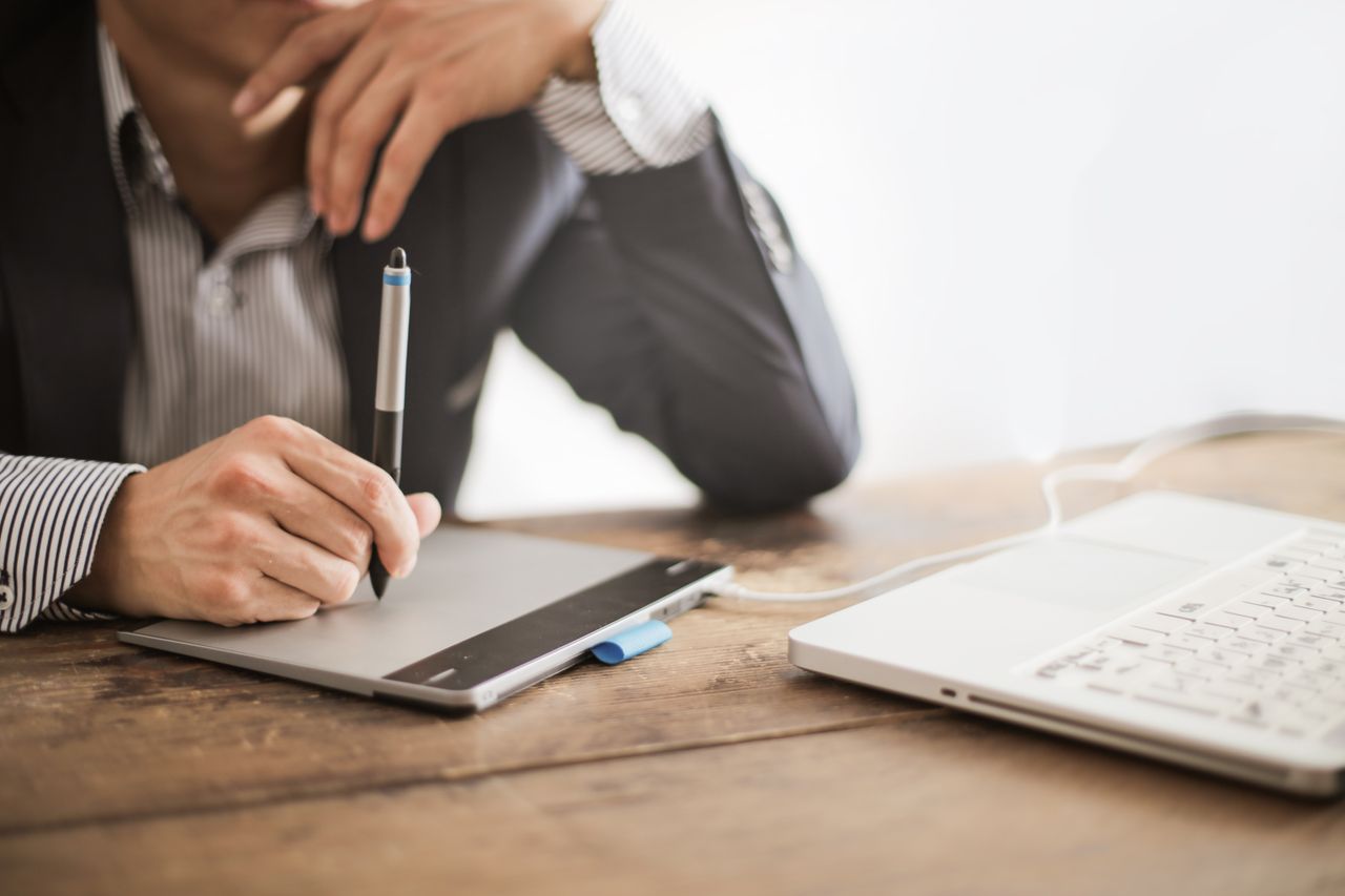 Offerings Crop elegant man watching laptop while drawing on graphic tablet at wooden desk